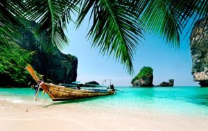 Gay beach Phuket TropOut: Longtail boat on a white sand beach with turquoise water and palm trees. Tropical gay travel destination.