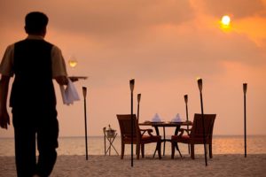 TropOut luxury dinner on beach at sunset. Waiter serving wine. Gay travel advice for romantic getaway.