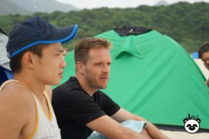 Gay Pride Hong Kong: Two men relax by a green tent on a beach during the Pink Season event. Mountains are visible in the background.
