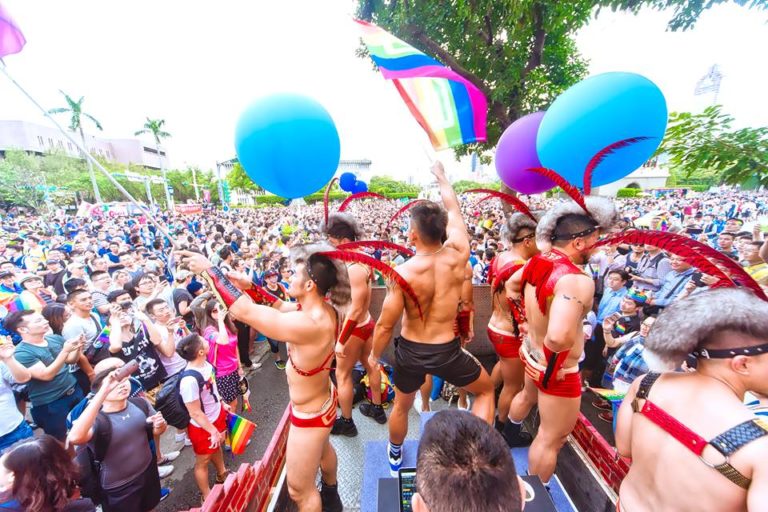 Taipei Pride 2017: Gay men in red costumes and feather headdresses dance on a float in front of a large crowd. Rainbow flag visible.