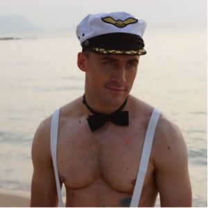 Gay beach in Pattaya, Thailand: Man with suspenders and bow tie, wearing a captain's hat on the beach. Gay travel destination.