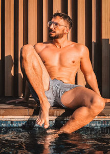 Shirtless man in swim trunks sits by a pool at X2 Chiang Mai Riverside Resort. Popular gay honeymoon destination for couples.
