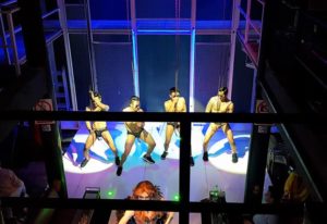 Jupiter 2018: Bangkok Go-Go Boys at Silom Soi 4. Four shirtless dancers in briefs hang from chains on stage. Gay nightlife scene.
