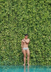 Gay Chiang Mai travel: Man at 137 Pillars House, one of the best gay hotels. Fit man in swim briefs by pool with green wall. TikTok.
