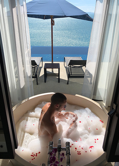 Man in bubble bath at Conrad Koh Samui, a gay-friendly hotel. Ocean view, deck chairs, and umbrella visible. Luxury travel for gay men.