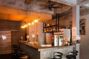 Bar area at The Beach Hostel, a gay-friendly hotel. Pendant lights hang above the counter with bar stools. Shelves of liquor behind.