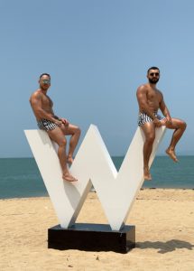 Two gay men in swim trunks pose on a W Koh Samui beachfront sculpture. Honeymoon private pool villa setting. Gay travel advice.