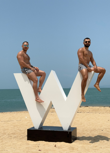 Two gay men in swim trunks pose on a W Koh Samui beachfront sculpture. Honeymoon private pool villa setting. Gay travel advice.