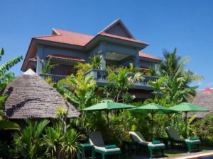 3 Monkeys Villa gay hotel. Exterior view of a gay-friendly, pet-friendly hotel with tropical foliage, lounge chairs, and thatched huts.