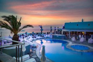 Elysium Hotel, a gay-friendly and pet-friendly hotel. Poolside view at sunset with people relaxing on white lounge seating by the sea.
