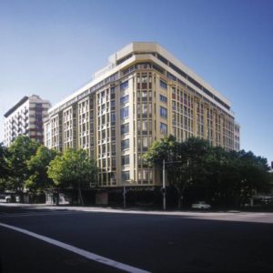 Exterior of Vibe Hotel Sydney, a gay-friendly hotel. The building is tall and modern, with many windows. Trees line the street.