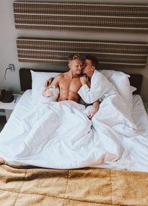 Gay couple in bed at Albus Amsterdam Centre, a boutique hotel. Man in robe kisses shirtless partner. Best gay hotels Amsterdam city center.