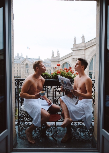 Gay honeymoon at 25hours Hotel Terminus Nord, Paris. Two men in towels on a balcony with coffee and newspaper, enjoying the city view.