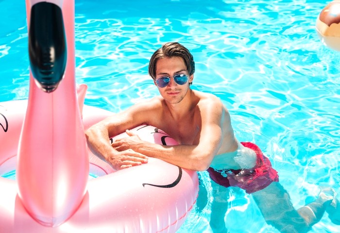 Man in a flamingo swim ring at a gay hotel in Sydney. Front view of a shirtless man with sunglasses in a pool. Gay travel.