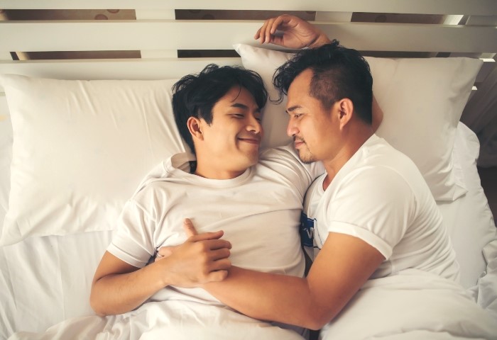 Gay couple love time in bed at a Hanoi hotel. Two men in white shirts lying down, smiling at each other. Gay travel.