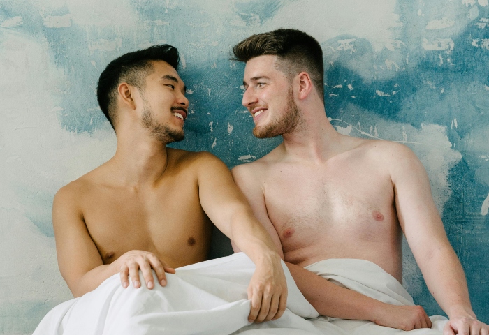 Gay couple smiling in bed at a Ho Chi Minh hotel. Two shirtless men relax under white sheets. Gay travel in Vietnam. Pexels 4670931.