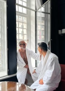 Gay honeymoon at The Dilly, a top gay hotel in London's Soho. Two men in robes smile near a window. Best gay honeymoon hotels.