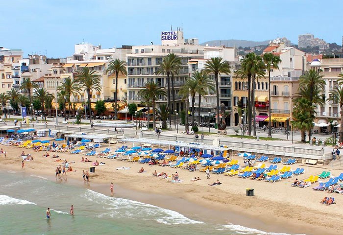 Hotel Subur Sitges, a gay-friendly and pet-friendly hotel. Beach view with sunbathers, palm trees, and the hotel in the background. Sitges, Spain.