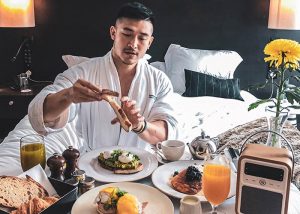 Gay man enjoys breakfast in bed at South Place Hotel, a pet-friendly hotel. He's wearing a robe and has toast, eggs, and juice.