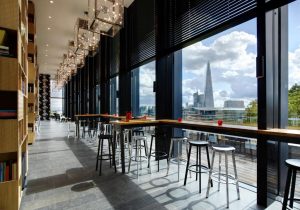 citizenM Tower of London's stylish interior. Bar stools line windows with city views. A gay-friendly hotel choice for your London travel.