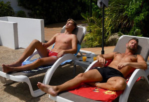 Two shirtless men relax on lounge chairs at Paso Chico Gay Hotel, Gran Canaria. Enjoying the sun at this gay party hotel in Yumbo Center.