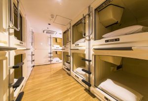 Interior of Shinjuku Kuyakusho-mae Capsule Hotel, a gay-friendly pod hotel. Rows of sleeping capsules with ladders and bedding are visible.