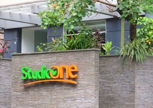 Studio One Hotel exterior. Gay-friendly hotel with green and orange logo on a textured wall. Lush plants above. Modern design.