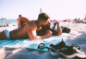 Gay Ibiza travel: Two men sunbathing on a beach. One man is wearing a white bandana and sunglasses, the other is shirtless and wearing swim trunks.