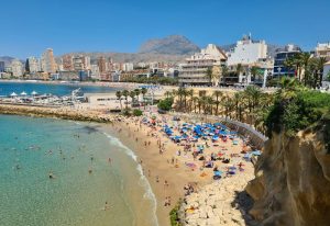 Benidorm beach view. Crowded sand with sunbathers, blue umbrellas, and clear water. Hotels and mountains in background. Gay travel destination.