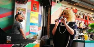 Dalston Superstore gay bar: Drag queen performing with a microphone. Man at the bar. Colorful wall art. Gay nightlife scene.