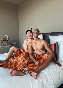 Gay couple in Berlin's Das Stue hotel. Two shirtless men cuddle under a blanket in a luxury hotel room near gay bars.