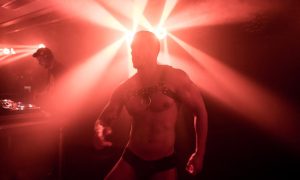 XLSIOR Mykonos gay party: shirtless man in harness dancing at a club with red lighting, DJ in the background. Best gay party in Mykonos.