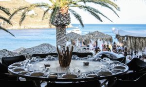 JackieO' Mykonos gay beach bar. Set table at gay bar overlooking the beach. Palm tree and ocean view. Mykonos gay travel.