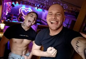 Two gay men having fun at a piano bar in Prague. The man on the left is shirtless. Heart shaped lights in the background.