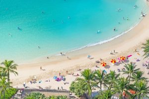 Aerial view of Waikiki Beach Tower, a gay-friendly hotel. Turquoise water, sandy beach, palm trees, and people swimming and sunbathing.