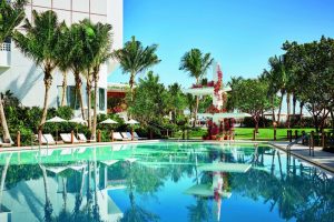Miami Beach EDITION's pool. Gay-friendly hotel with palm trees, lounge chairs, and a serene pool reflecting the blue sky. A tropical paradise.