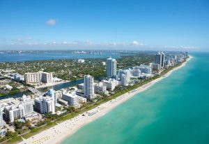 Aerial view of Miami Beach, a top gay travel destination. Turquoise ocean meets the sandy shore, lined with hotels and condos under a clear blue sky.