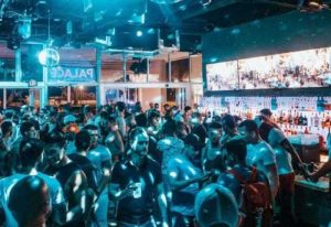 Packed Palace Bar, a popular gay bar. Men gather under disco lights, with a fully stocked bar visible in the background. Nightlife scene.