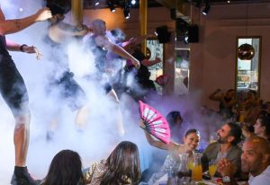 Gay men's travel: R House's drag brunch. Dancers perform amidst smoke, with guests cheering and waving fans. 5-19.jpg captures the vibrant scene.