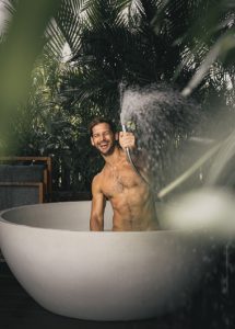 Gay honeymoon: Man in tub at Nirjhara Bali, a luxury hotel. He's smiling, holding a showerhead, surrounded by lush greenery. Best gay hotel Bali.