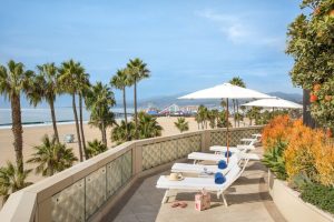 Casa Del Mar gay-friendly hotel view. Beachfront with palm trees, lounge chairs, umbrellas. Santa Monica Pier visible. Perfect gay travel spot.