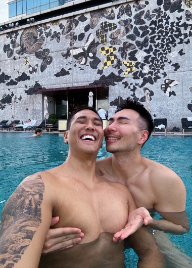 Gay honeymoon at W Hong Kong hotel pool. Two shirtless men embrace, smiling, with butterfly mural backdrop. Perfect gay travel destination.