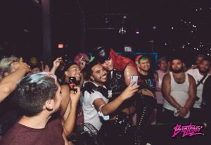 Precinct Party: Gay men at a party take a selfie with a drag queen in fetish gear. People are holding up their phones to take pictures.