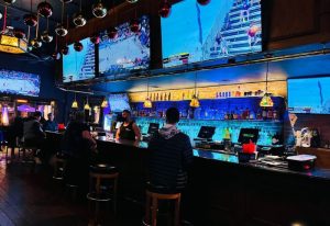 Interior of TRUNKS Bar, a gay bar. People sit at the bar watching sports on large screens. Blue lighting and disco balls create a vibrant atmosphere.