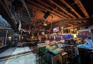 Interior of The Abbey Bar, a gay bar. The bar area is visible, with bottles and glasses. The ceiling has exposed wooden beams and lighting fixtures.