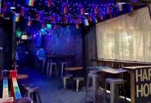 The Bayou dancefloor: Gay bar interior with rainbow flags, blue lighting, tables, and stools. Happy hour sign. Gay travel destination.