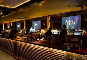 The Chapel @ The Abbey bar interior. Bartenders serve drinks at a long bar. Screens display The Chapel logo. Gay travel destination.