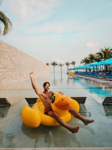 Gay Songkran travel: Man on inflatable duck in pool. Tropical resort with palm trees. Gay travel advice, fun vacation, a5732f99-eb96.