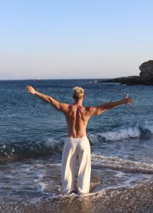 Akkadian Gay Only Hotel Mykonos: Man with arms outstretched on a beach. Best gay beach hotels in Mykonos for gay men's travel advice.