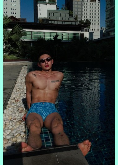 YanYiFan at Pullman Bangkok King Power pool. Gay travel in Thailand. Man in swim trunks, sunglasses, and lipstick relaxes poolside.